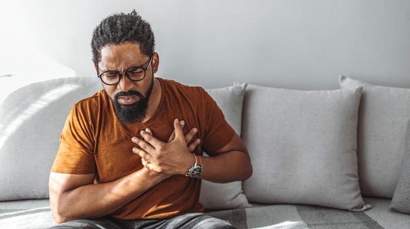 An African-American man clutches his chest while sitting on his livingroom sofa.