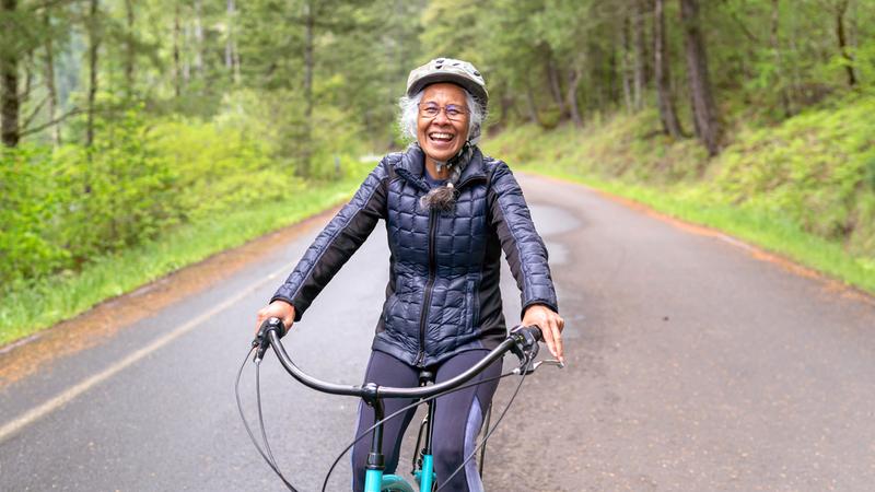 A senior woman rides a bicycle on a road in a wooded area. She is wearing a helmet and a warm coat.