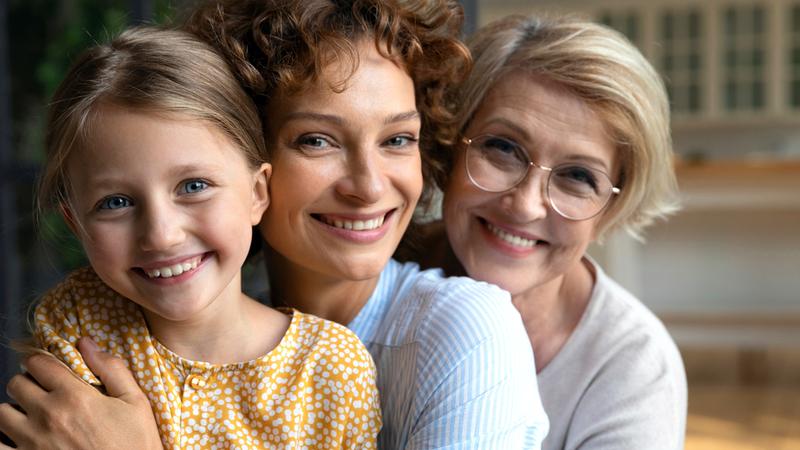 3 women, a girl, mother, and grandmother, pose for a photo in their home.