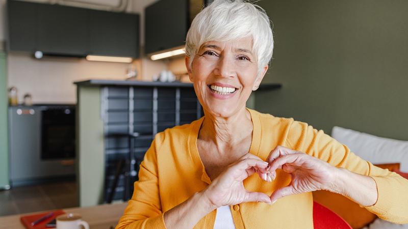 A senior woman smiles and looks at the camera as shesits in a chair in her kitchen and makes a heart symbol with her hands.